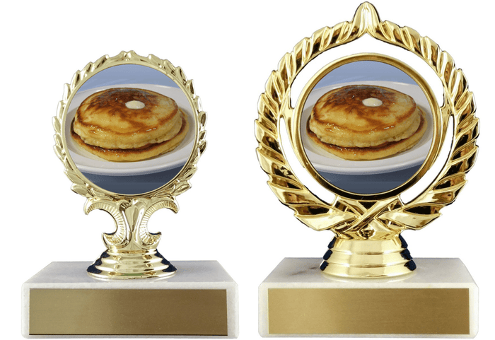 Two gold trophy cups with oval medallions showing a stack of pancakes, mounted on white bases with gold front plates, photographed on a white background.